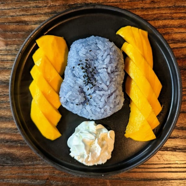 Mango sticky rice with blue rice, fresh mango slices, and a dollop of cream on a black plate.